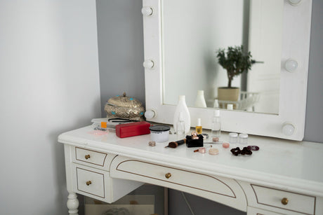 “Decorated dressing table with a mirror, makeup organizers, perfume bottles, and a small vase of flowers in a well-lit bedroom.”
