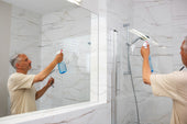 “Person cleaning a glass shower screen with a squeegee and spray cleaner to remove soap scum and water spots.”