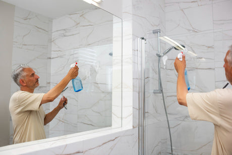 “Person cleaning a glass shower screen with a squeegee and spray cleaner to remove soap scum and water spots.”