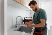 “Person installing a stainless steel kitchen sink into a countertop cutout, connecting the faucet and plumbing underneath.”